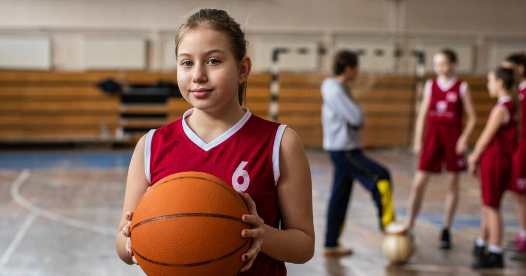 young girl with basketball standing in an indoor basketball court, basketball team and coach are in the background